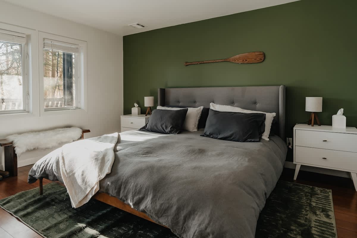 A serene bedroom featuring a large gray upholstered bed against a dark green accent wall, flanked by white mid-century modern nightstands with lamps. Hardwood floors are partially covered by a green area rug.