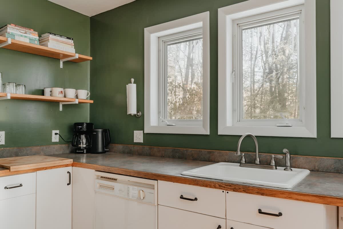 Bright kitchen corner featuring a white farmhouse sink with a gooseneck faucet, set into a butcher block countertop. Two windows above the sink offer views of the outdoors, and a coffee maker and kettle are visible on the counter.