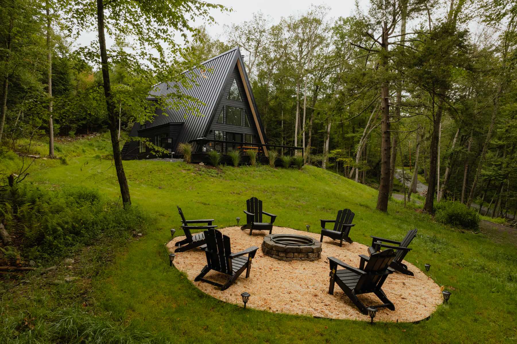 Outdoor fire pit area with a circular gravel base surrounded by black Adirondack chairs and small lights, set on a grassy hill with a modern black A-frame house and dense forest in the background.