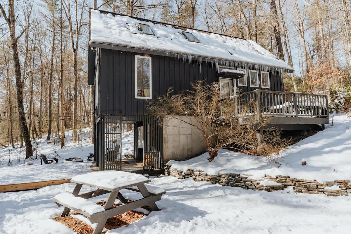 Wide exterior view of the dark gray house in a snowy winter landscape, featuring a wooden deck, a snow-covered picnic table, and icicles hanging from the roof. Bare trees surround the property, highlighting its secluded setting.