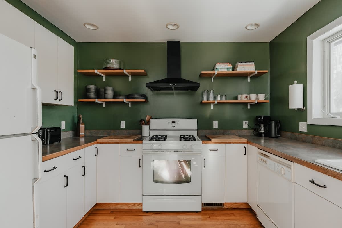 Modern kitchen with striking dark green accent walls and white shaker-style cabinets. A white gas range with a black range hood is centered, flanked by open wooden shelves and butcher block countertops.