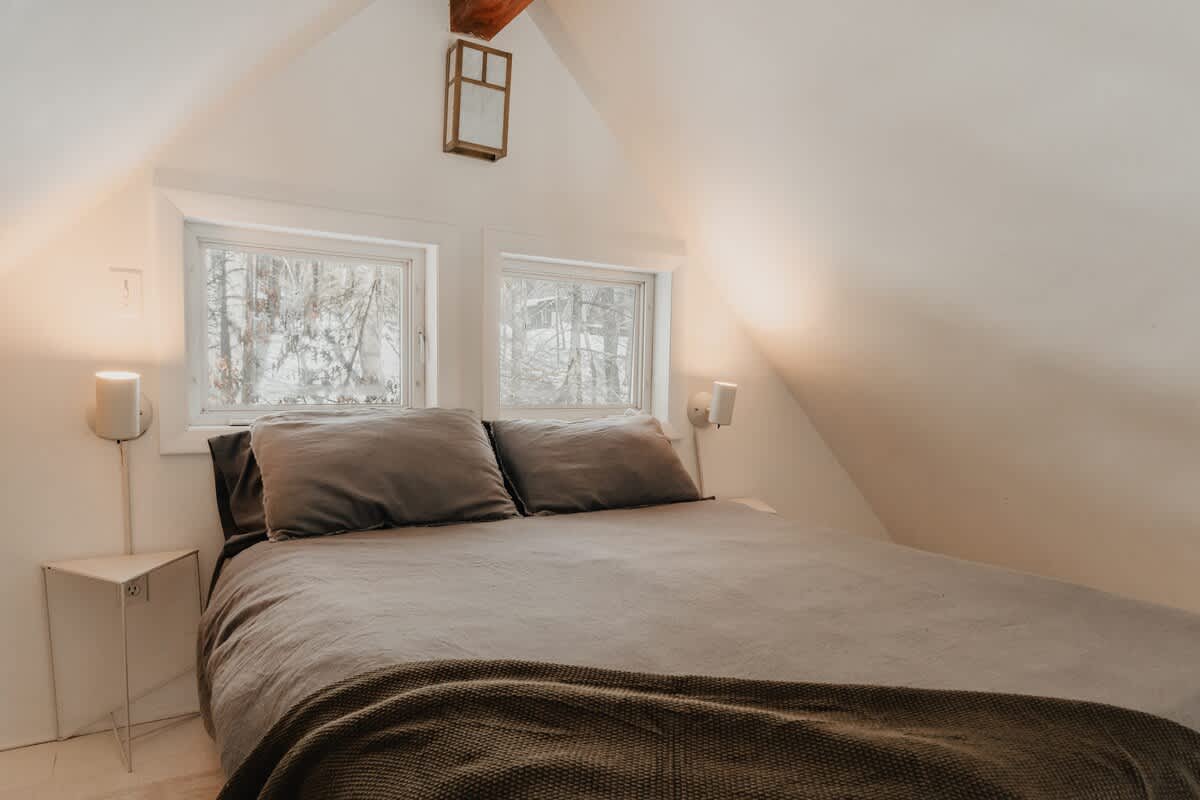 Cozy attic bedroom with a bed featuring a gray duvet and pillows, flanked by two small windows offering a view of snowy trees. Wall-mounted lamps provide soft lighting, and a wooden beam is visible in the vaulted ceiling.