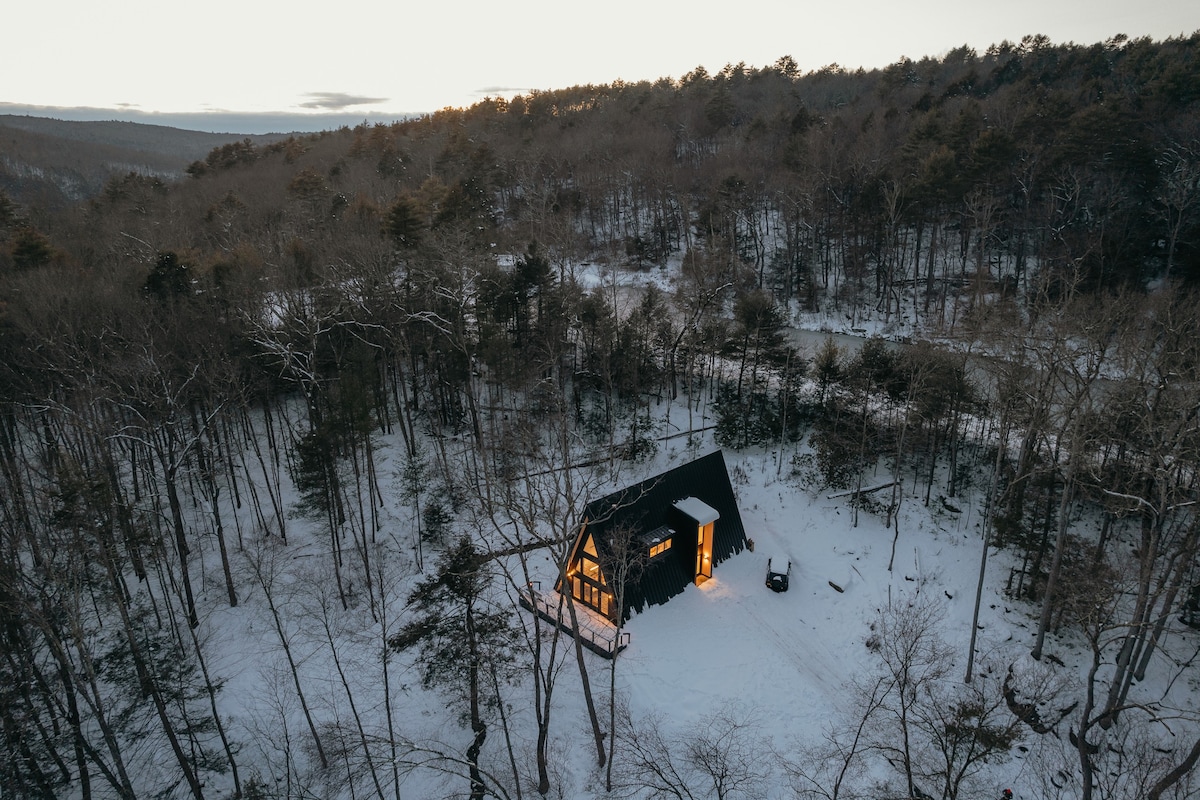 Aerial view of a modern black A-frame cabin nestled in a snow-covered forest, with warm lights glowing from its windows, surrounded by bare trees and distant hills under a soft, overcast sky.