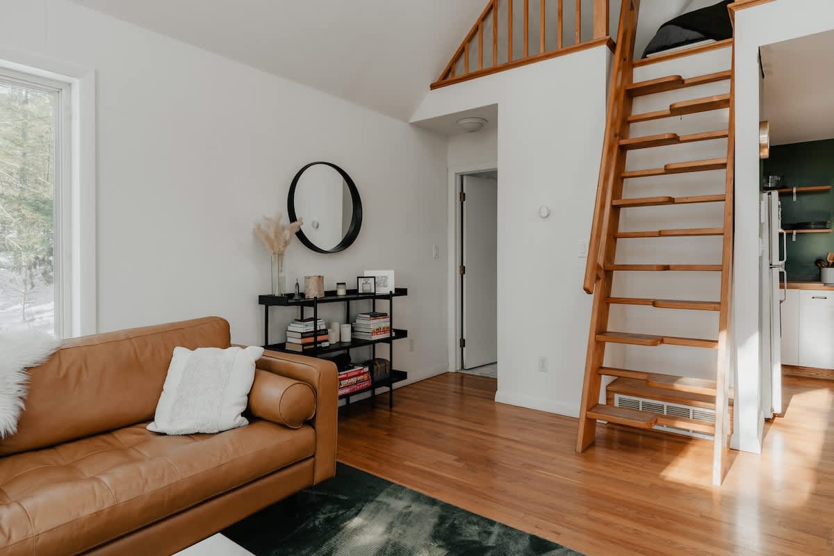 A bright and airy living room featuring a comfortable brown leather sofa, a black metal shelving unit filled with books and decor, and a round black mirror. A unique wooden ladder-style staircase leads to an upper level.