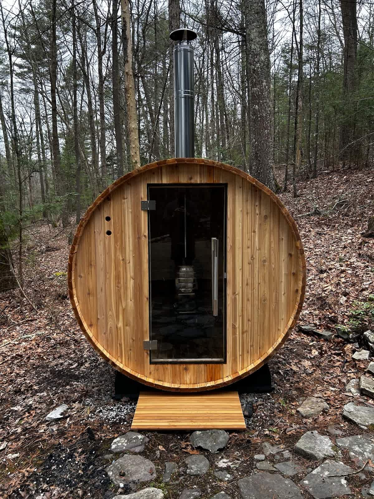 Cylindrical wooden barrel sauna with a glass door and a metal chimney, set amidst a forest floor with rocks and fallen leaves. The interior shows a wood-burning stove.