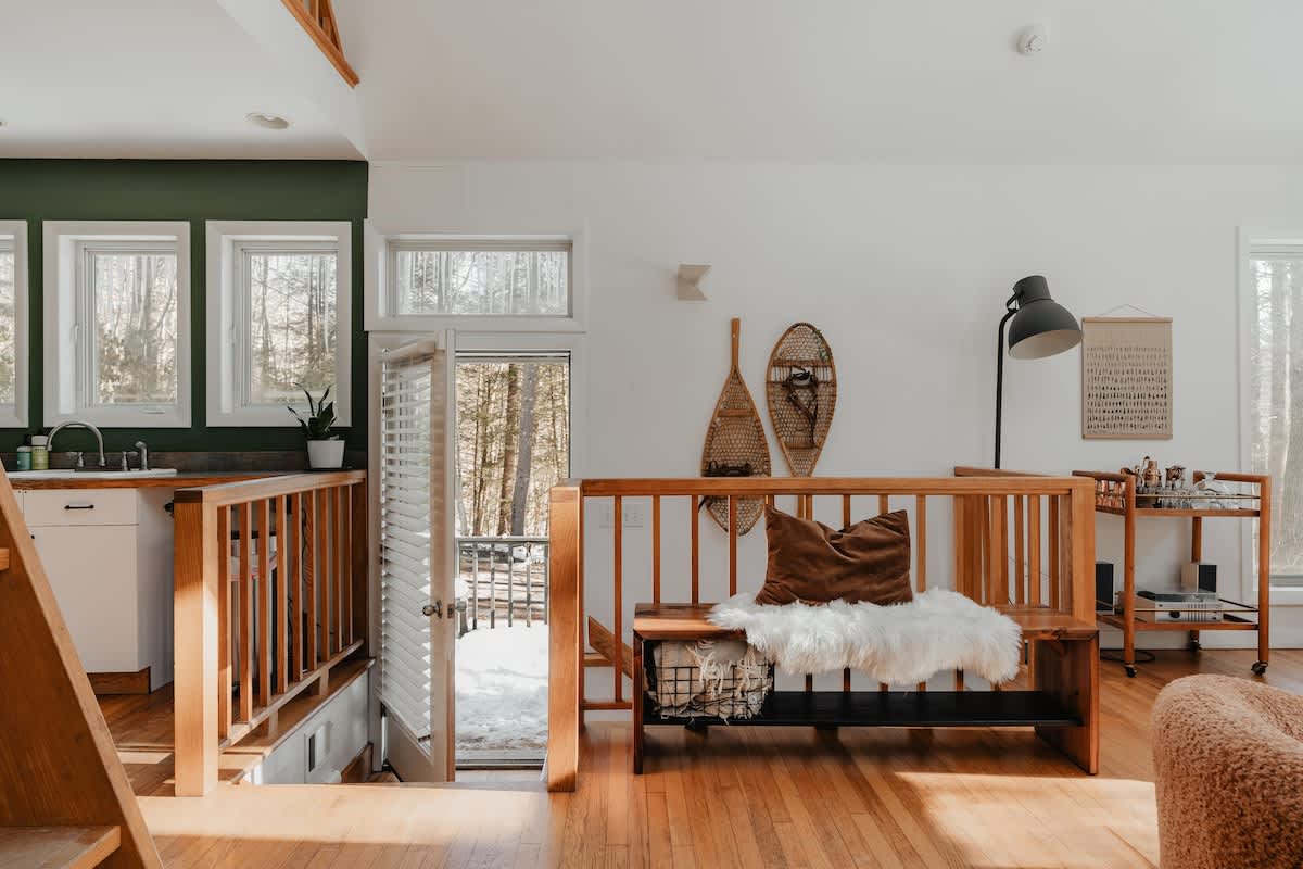 View of the living area from a raised perspective, showing a wooden bench with a faux fur throw and decorative snowshoes on the wall. An open doorway leads to an outdoor deck, and a bar cart is visible to the right.