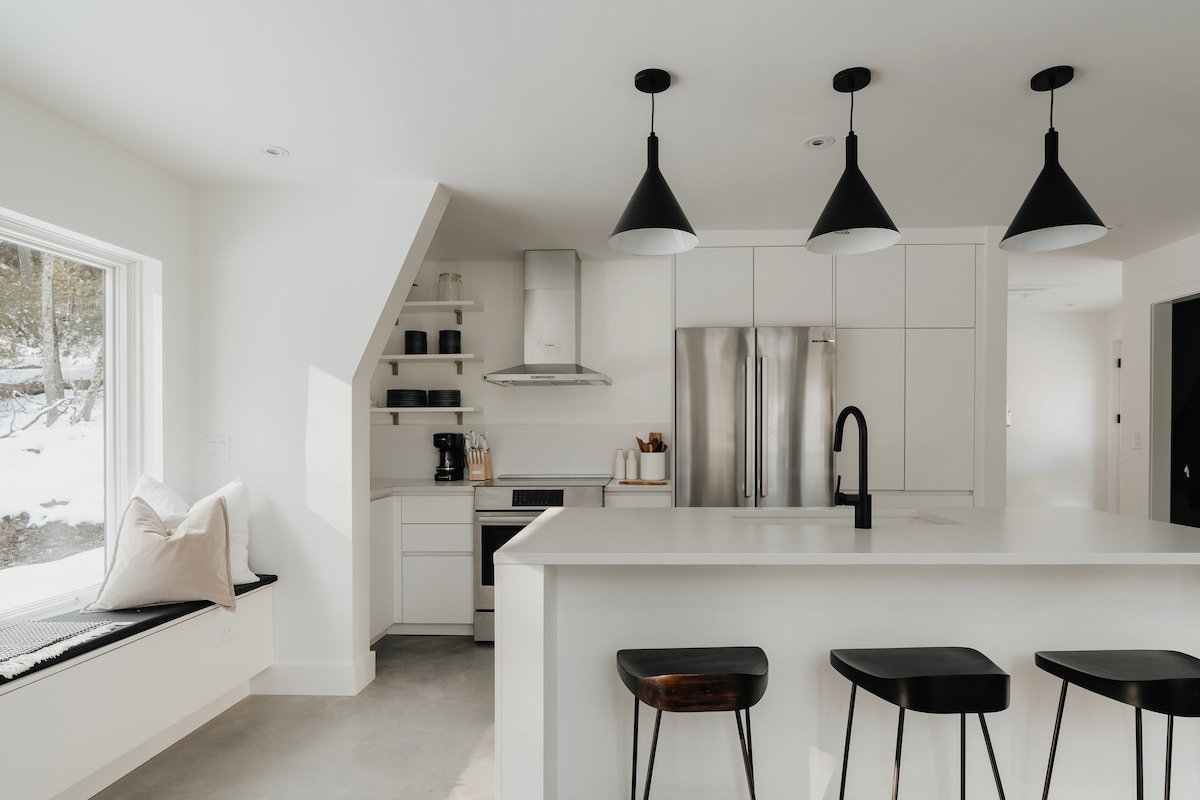 Modern white kitchen featuring a large island with bar stools, stainless steel refrigerator, and a range hood. Open shelving and a window seat with pillows add functional and cozy elements to the bright space.
