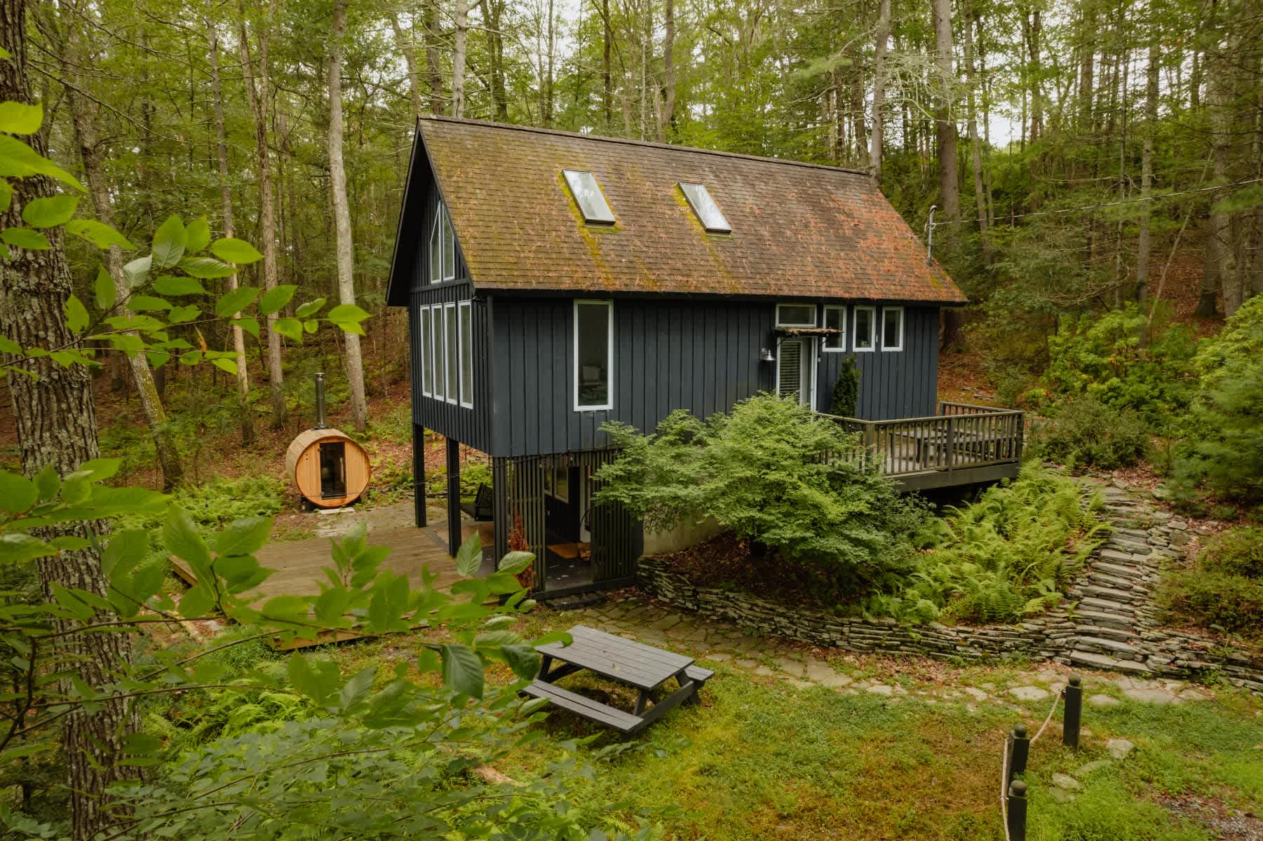 A dark gray A-frame house with a brown shingled roof and skylights, nestled among lush green trees. A barrel sauna sits on a wooden deck to the left, and a picnic table is visible in the grassy foreground.