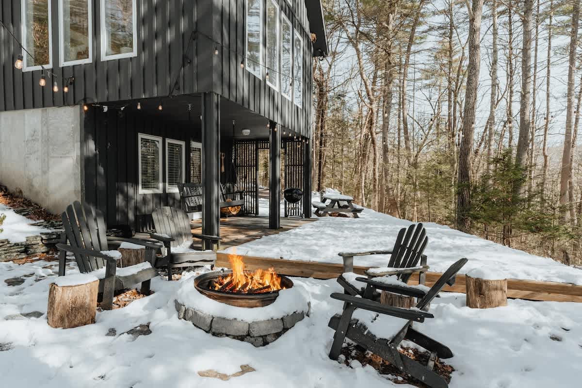 Winter scene of an outdoor fire pit area, surrounded by snow-covered Adirondack chairs and tree stumps. A warm fire burns in the stone-ringed pit, with the dark house and bare trees in the background.