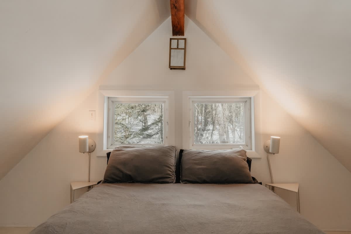 Symmetrical view of a cozy attic bedroom with a bed featuring a gray duvet and pillows, centered between two small windows looking out to snowy trees. Wall-mounted lamps illuminate the sloped white walls.