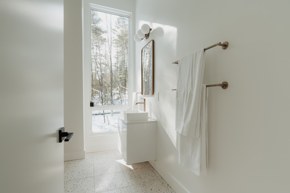 Bright, modern bathroom with white walls, a square vessel sink on a white vanity, a rectangular mirror, and a large window overlooking a snowy forest, set on a light terrazzo floor.