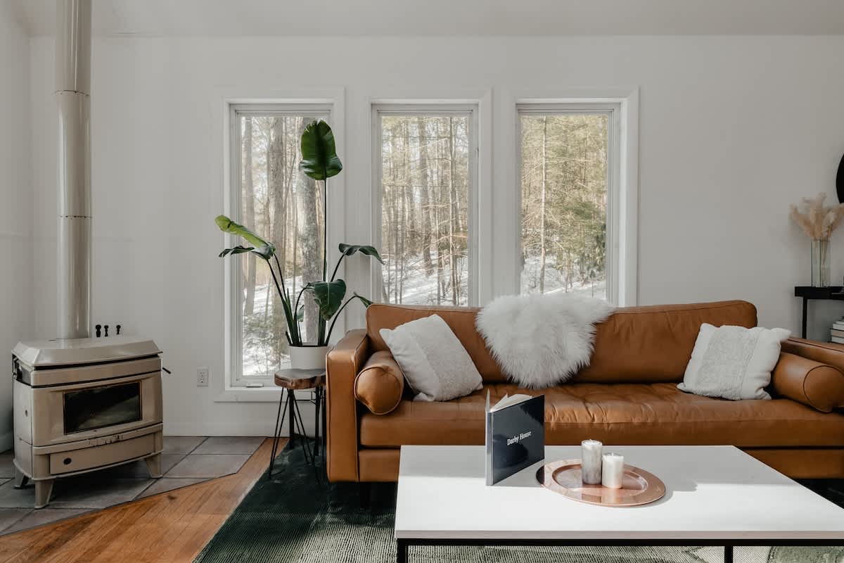 Cozy corner of the living room with a tan leather sofa adorned with white pillows and a faux fur throw. A modern white coffee table sits on a dark green rug, facing a cream-colored wood-burning stove with a tall metal flue.