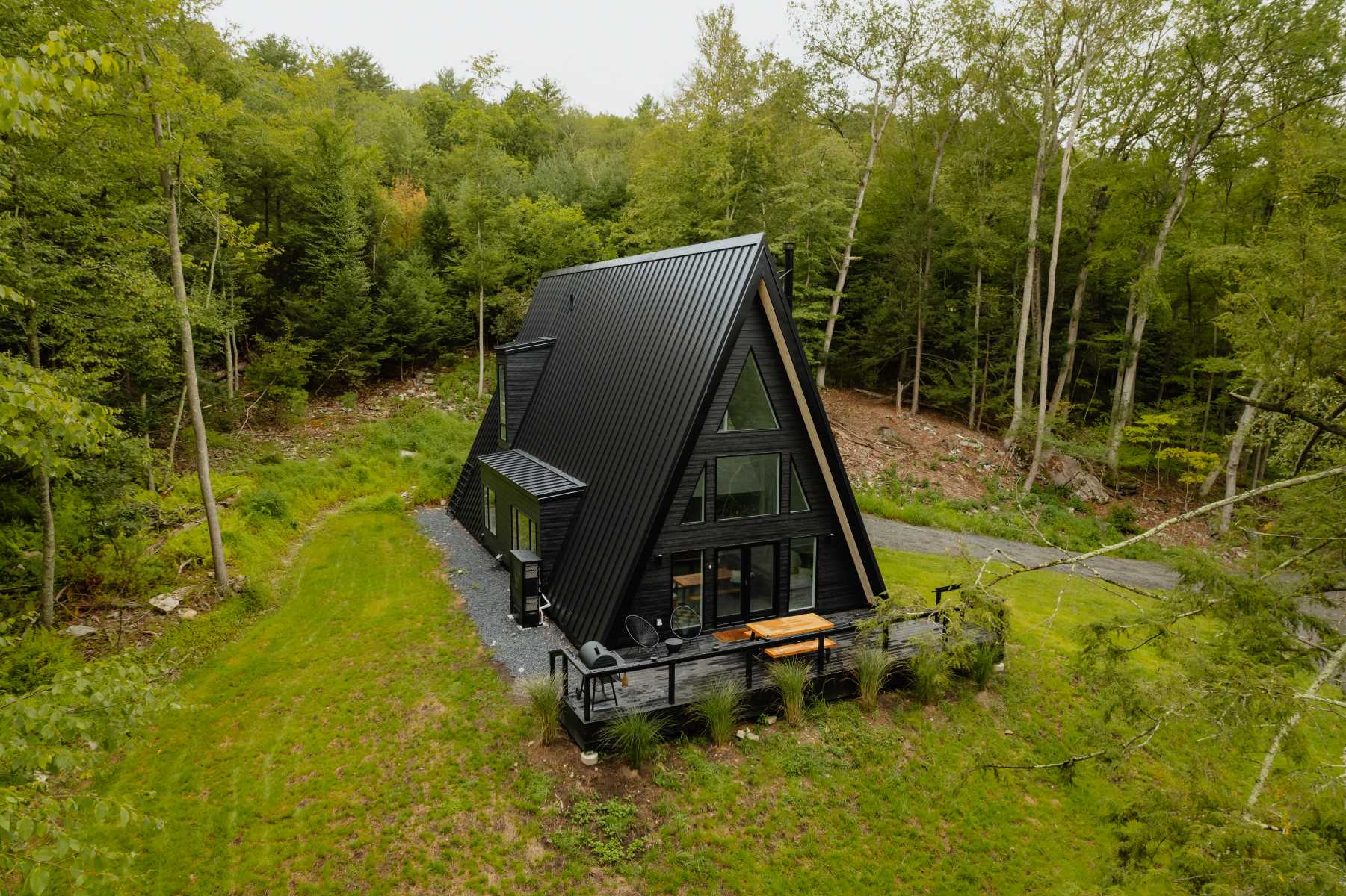 Aerial view of the black A-frame cabin nestled in a lush green forest, showcasing its steep roof and a spacious deck. A gravel driveway leads to the house, surrounded by natural landscaping.