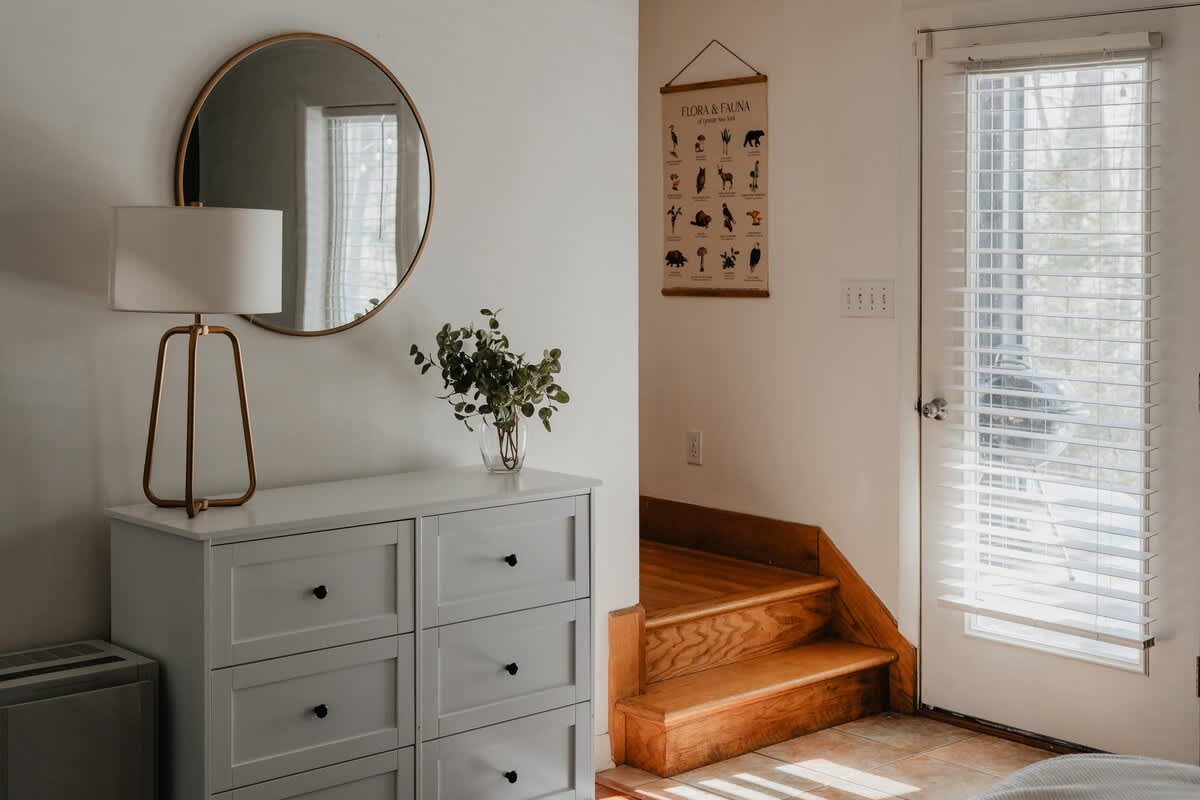 A bright entryway with a light gray dresser topped with a round gold-framed mirror and a vase of greenery. Wooden stairs lead upwards, adjacent to a door with white blinds, on a tiled floor.