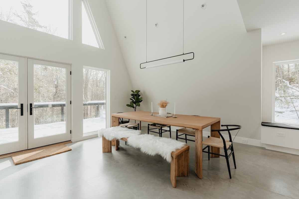 Spacious dining area featuring a long light wood table with a matching bench covered in white faux fur throws, and black chairs. Large windows overlook a snowy landscape, complemented by a modern linear pendant light and a potted plant.