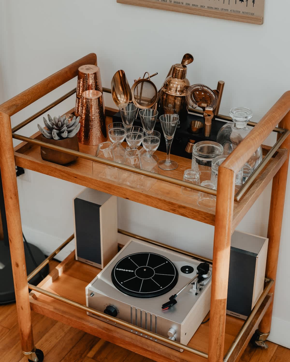 Close-up of a stylish wooden bar cart on wheels, stocked with various cocktail tools, glassware, and a vintage-style record player with speakers on the bottom shelf.