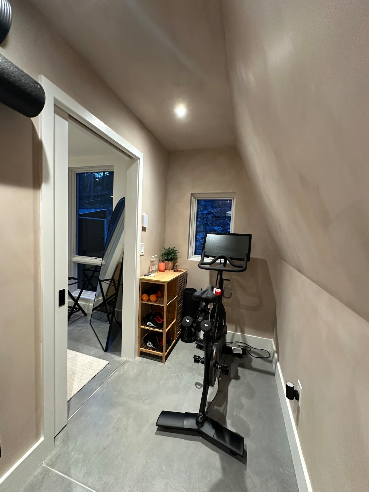 Compact exercise room with a black Peloton bike, a small wooden shelving unit, and a high window, featuring sloped ceilings and light-colored walls over a concrete floor.