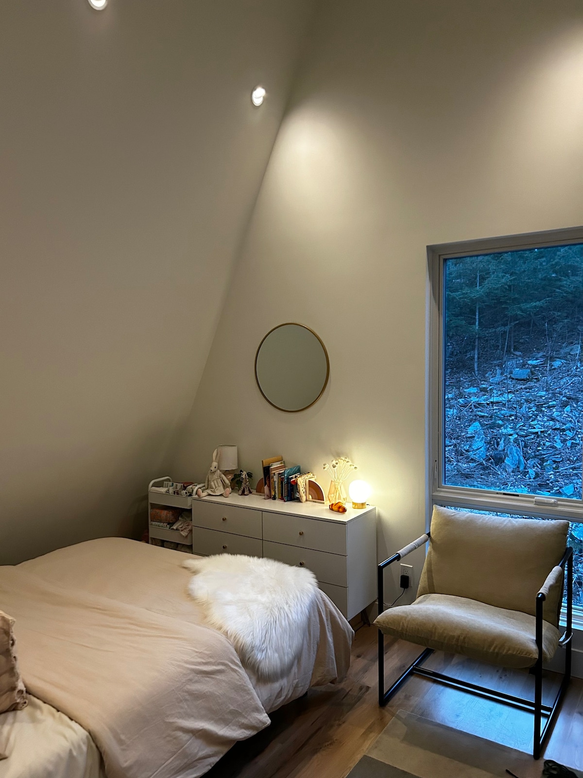 Cozy bedroom with a sloped ceiling, a bed with light peach bedding, a white dresser with a round gold mirror, and a comfortable beige armchair next to a large window with a dark forest view.