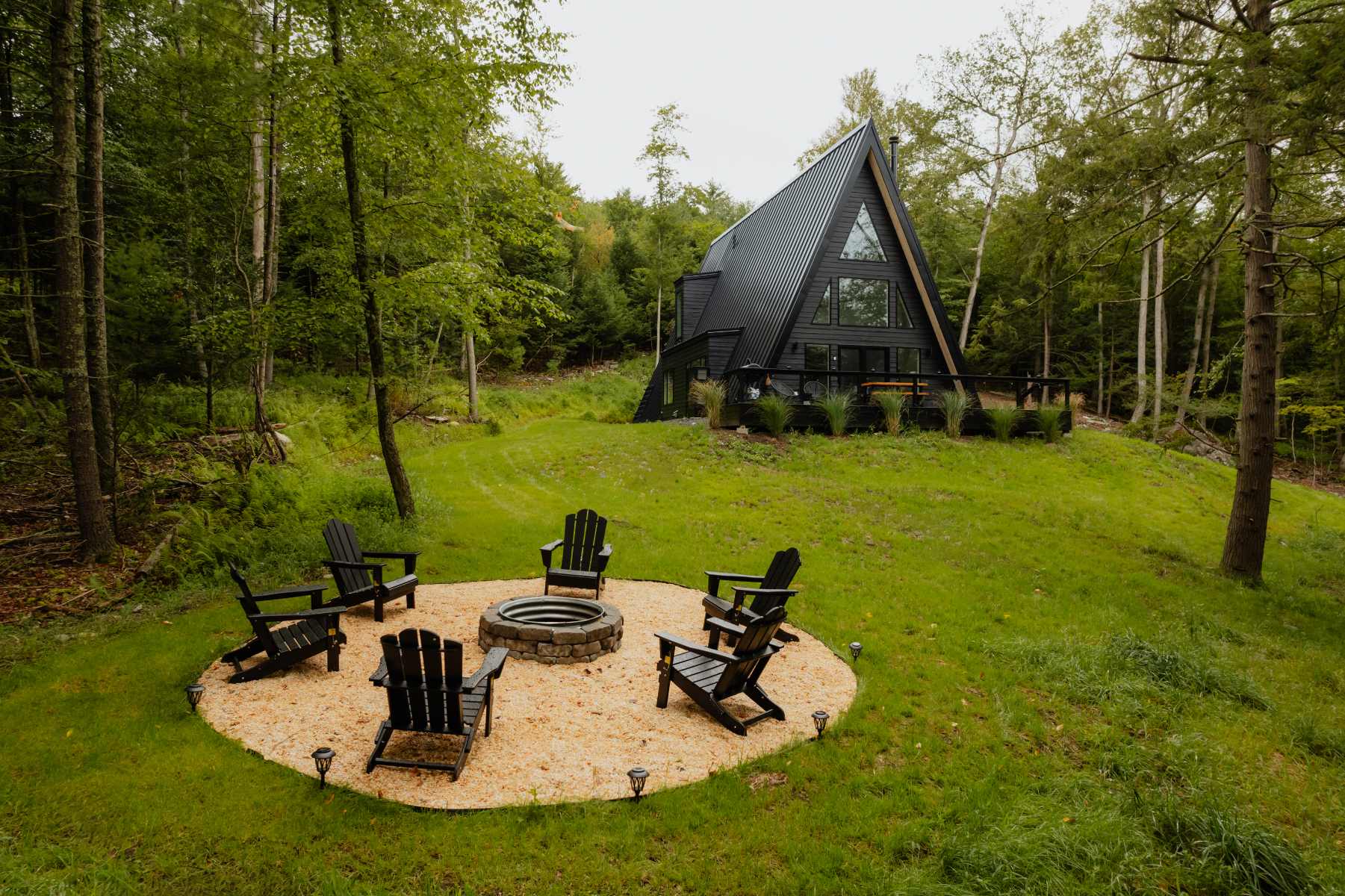 Outdoor fire pit area with a stone ring, surrounded by six black Adirondack chairs on a gravel base, set in a grassy clearing with the black A-frame cabin visible in the background among green trees.