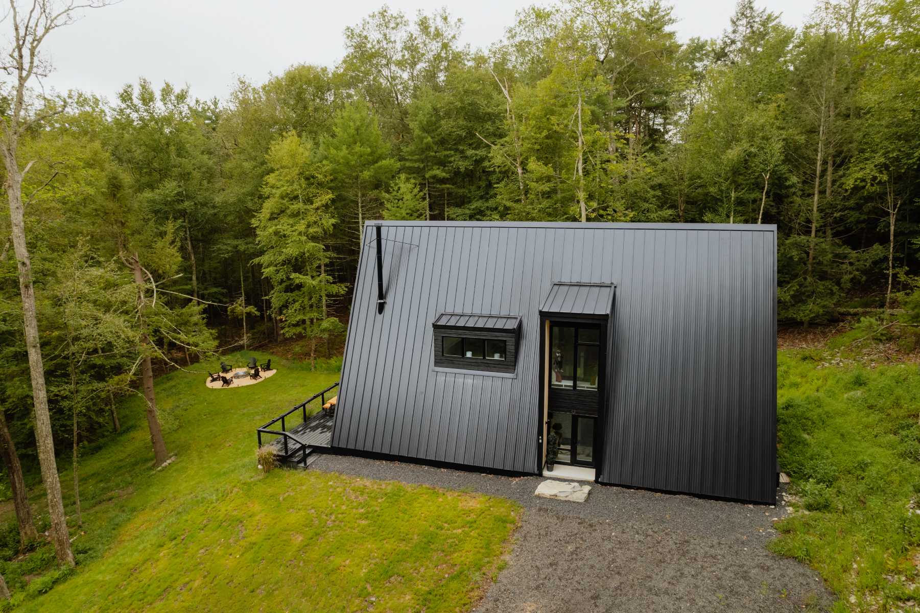 Aerial view of a modern black A-frame house with a corrugated metal roof, situated on a green hill surrounded by a lush forest, with a fire pit area and gravel driveway visible in the distance.