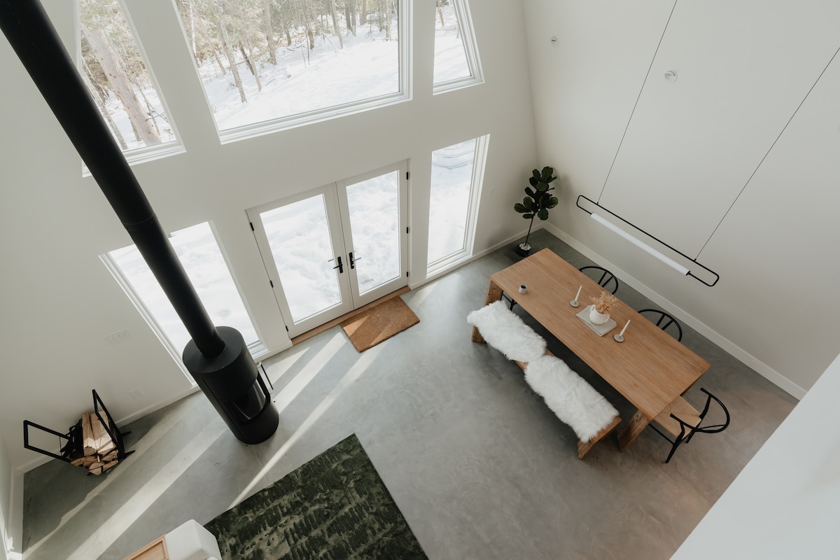 An aerial view showcases the open-concept living and dining areas with soaring ceilings and large windows overlooking a snowy landscape. A modern black fireplace anchors the living space, adjacent to a white sofa and a wooden dining table.