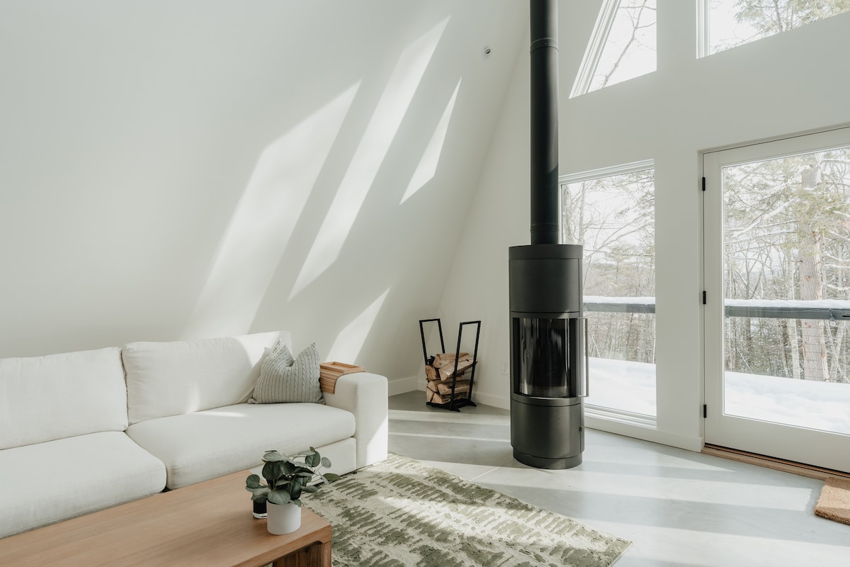 Bright living room with a modern black cylindrical fireplace, a white sofa, and large windows offering a snowy forest view. Sunlight streams across the concrete floor, highlighting a green patterned rug and a wooden coffee table.