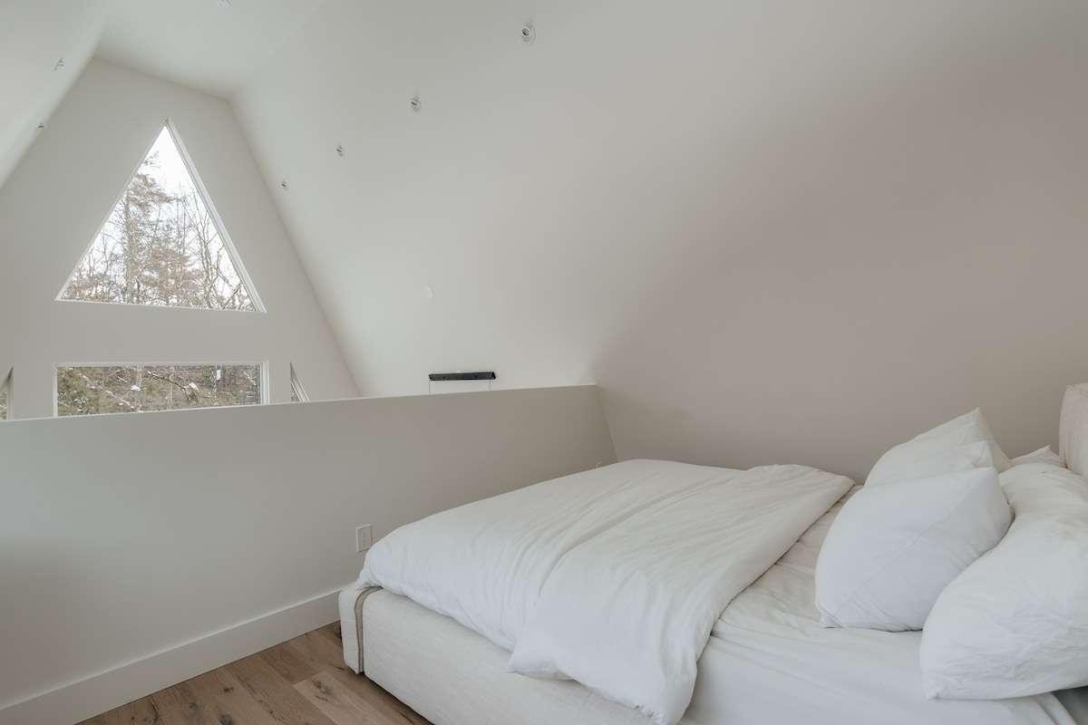 Minimalist bedroom with a low-profile white bed and crisp white bedding, set against white walls. A unique triangular window and additional rectangular windows offer views of the snowy outdoors, with light wood flooring.