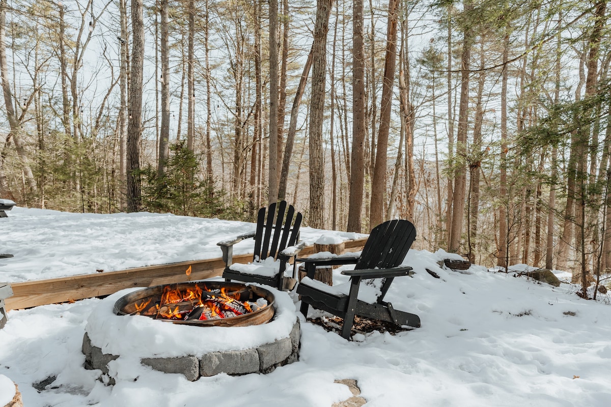 A close-up of the stone fire pit with a bright fire burning, surrounded by snow-covered ground and dark Adirondack chairs, set against a backdrop of tall, bare trees.