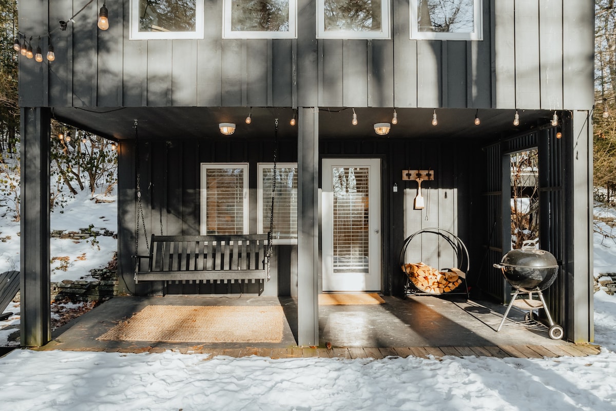 The covered porch area of the cabin in winter, featuring a dark gray porch swing, a white entry door, and a charcoal grill, with string lights overhead and snow on the ground.