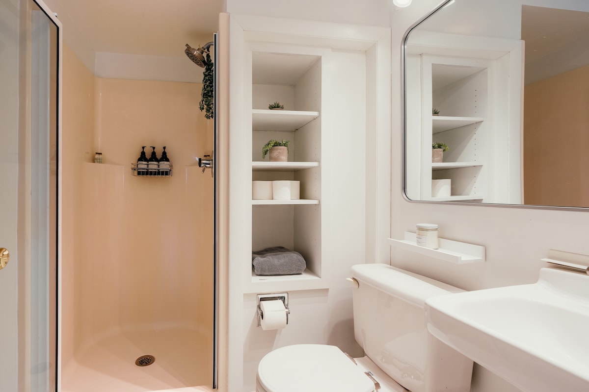 A compact bathroom features a white toilet and sink vanity, with built-in shelving next to the toilet holding toiletries and decorative plants. A peach-colored shower stall with a rainfall showerhead is visible on the left.