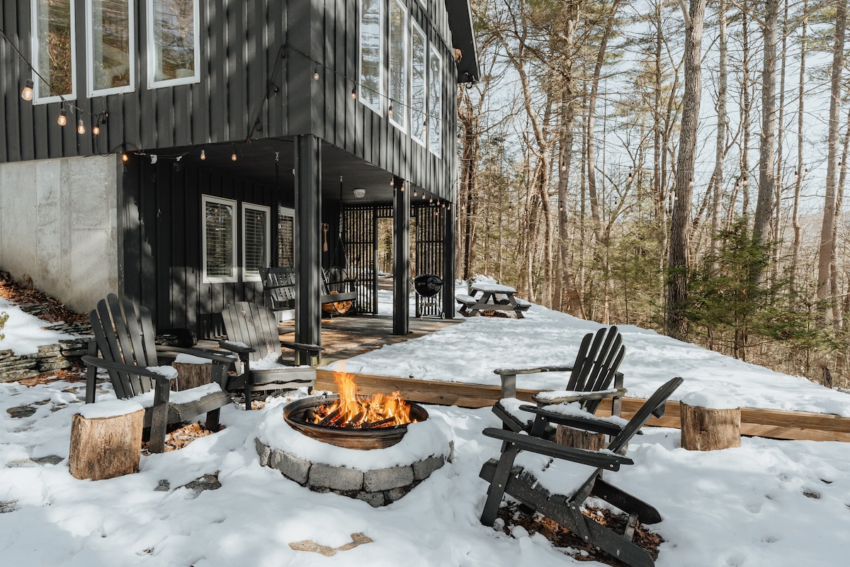 A cozy outdoor fire pit area in winter, surrounded by snow-covered ground, featuring a stone fire pit with a roaring fire and several dark Adirondack chairs on tree stump side tables.