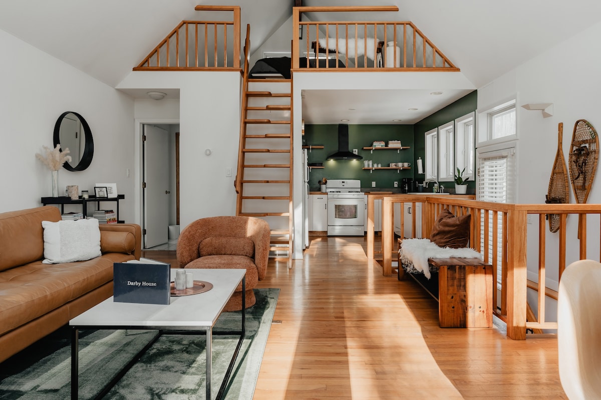 Open-concept living area with hardwood floors, a brown leather sofa, a dining table, and a view into the kitchen with a dark green accent wall and open shelving. Loft stairs are visible.