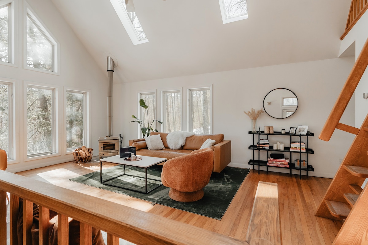 Bright, open-concept living room with a high vaulted ceiling, skylights, and large windows offering forest views. It features a brown leather sofa, a round orange armchair, a black coffee table on a dark green rug, and a wood-burning stove in the corner.