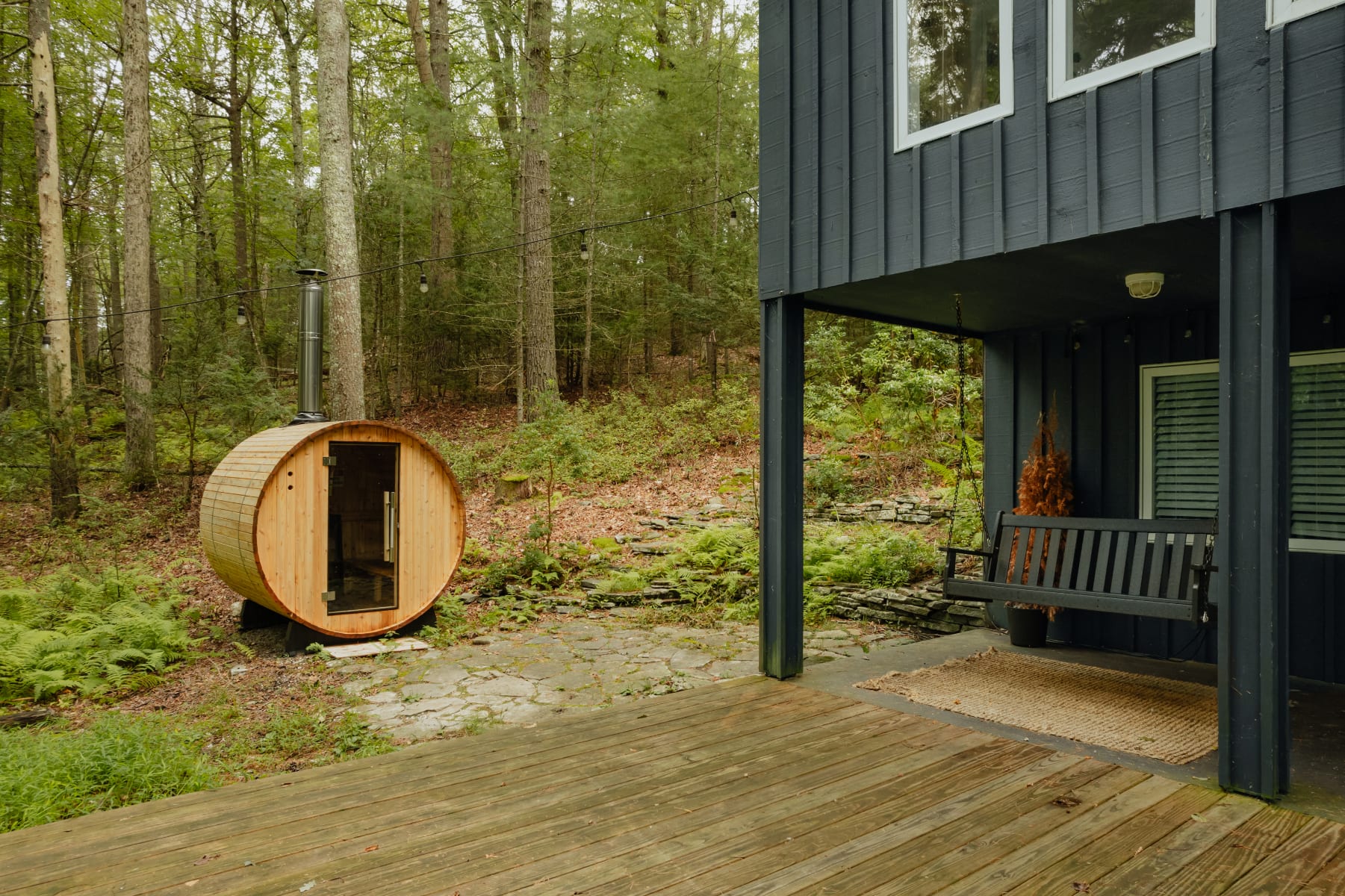 The covered porch area with a dark gray swing, leading to a stone patio and a distinctive wooden barrel sauna with a glass door, surrounded by a lush forest.