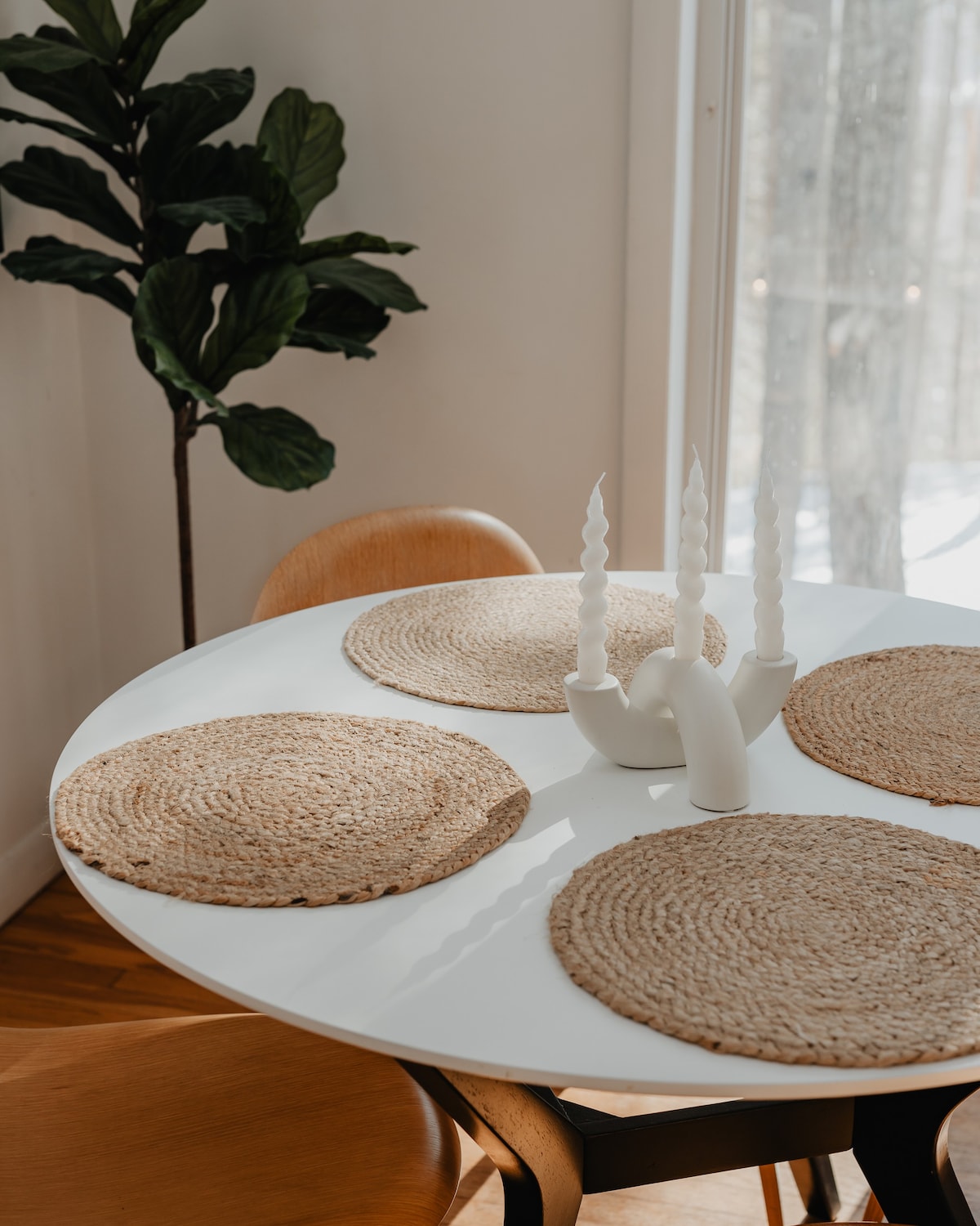 Close-up of the round white dining table set with four woven placemats and a modern white candelabra holding twisted white candles. A large green houseplant is visible in the background, adding a touch of nature.