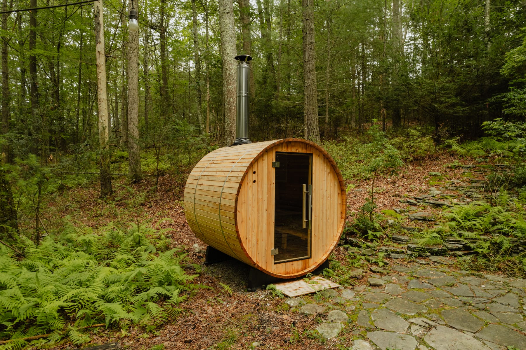 A close-up of the wooden barrel sauna with a glass door and a metal chimney, situated on a stone path amidst green ferns and tall trees in a forest setting.