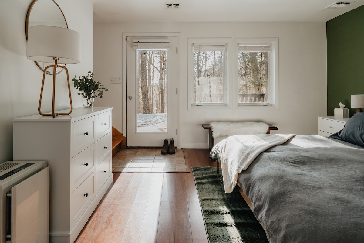 A bright bedroom with white walls and hardwood floors, featuring a large bed with gray bedding and a white throw, and a wooden bench with a sheepskin throw under two windows. A glass-paned door leads outside, revealing a snowy landscape.