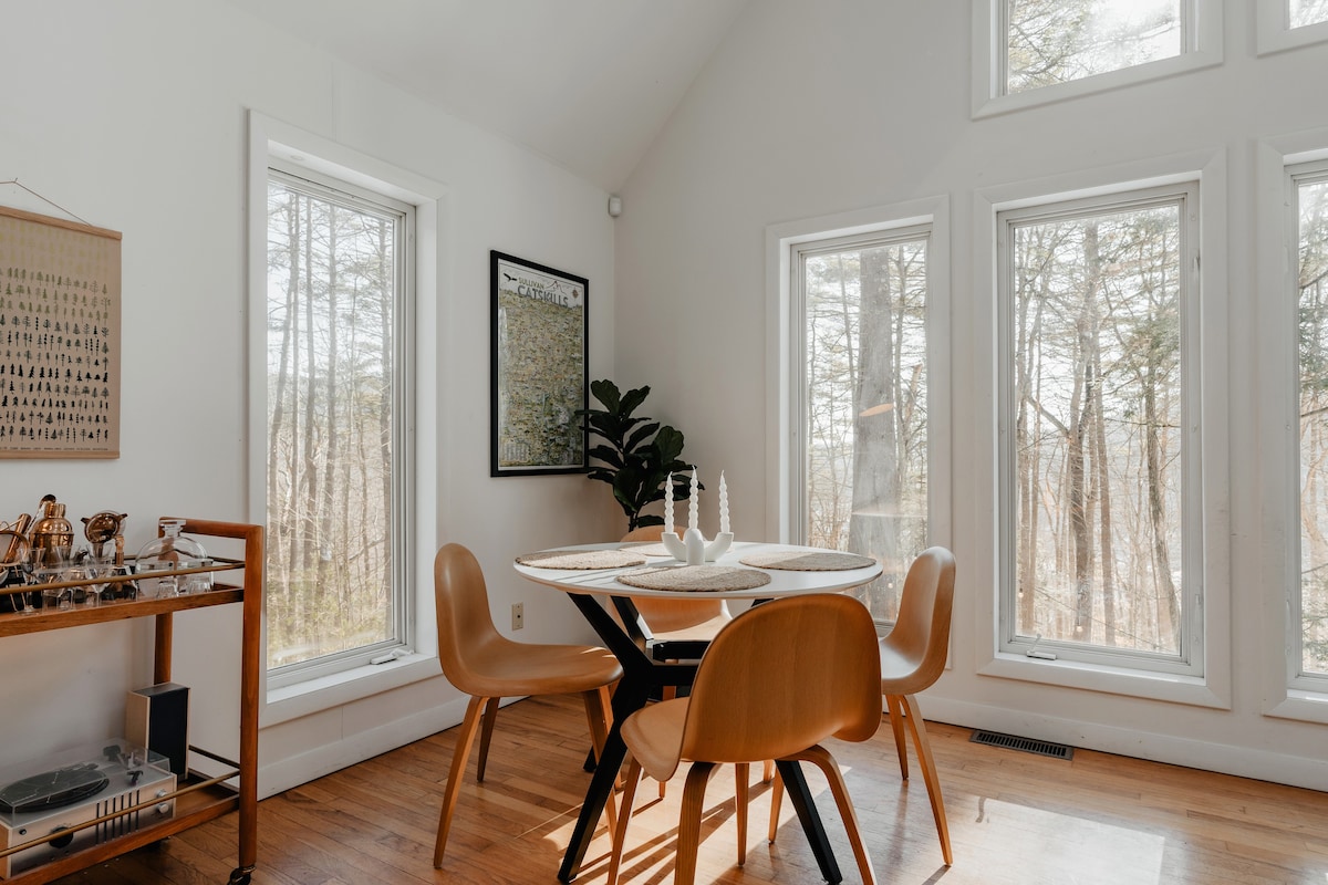 Dining area with a round white table, four light brown chairs, and large windows overlooking a winter forest. A framed map hangs on the wall, and a wooden bar cart with glassware is visible in the background.
