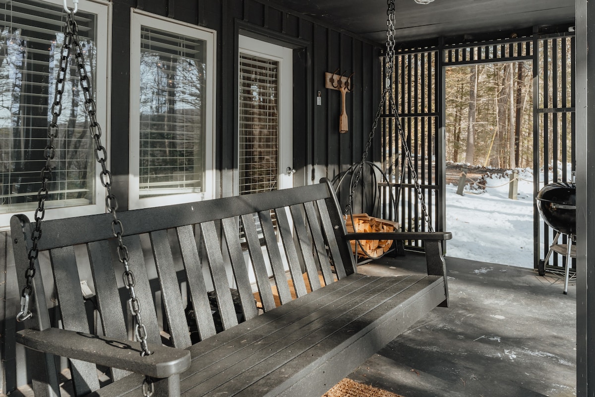 A close-up view of the dark gray wooden porch swing on the covered porch, with snow visible outside, and string lights adding a warm ambiance.