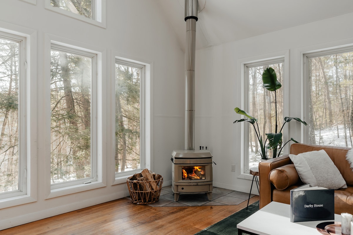 Close-up of the living room corner featuring a cream-colored wood-burning stove with a visible fire, set on a tiled hearth. Large windows surround the stove, showcasing a snowy forest outside, and a brown leather sofa is partially visible.