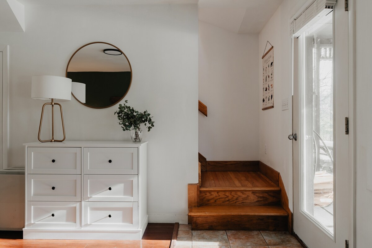 A well-lit entryway showcases a white dresser with a lamp and plant, a large round mirror, and wooden stairs leading upwards. The space transitions from hardwood to tiled flooring near the glass-paned door, which has a "Flora & Fauna" poster next to it.