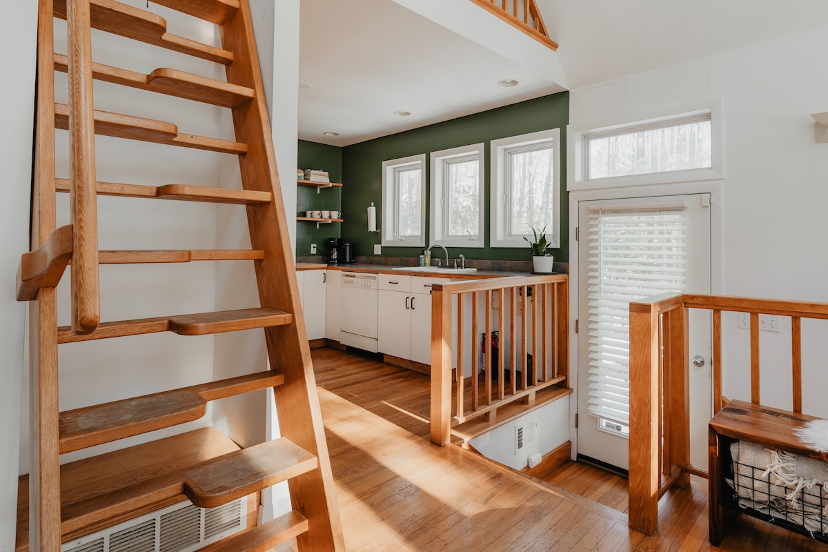 View of the kitchen area with white cabinets and a dark green accent wall, seen from the top of a wooden staircase. A door with blinds leads to the outside, and a wooden railing separates the kitchen from the lower level.