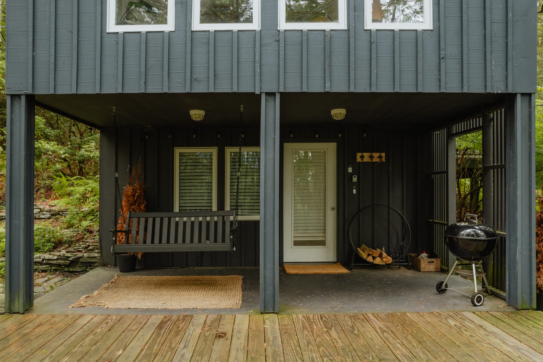 A covered porch area beneath the cabin, featuring a dark gray porch swing, a white entry door, a charcoal grill, and stacked firewood, all set against the dark vertical siding.