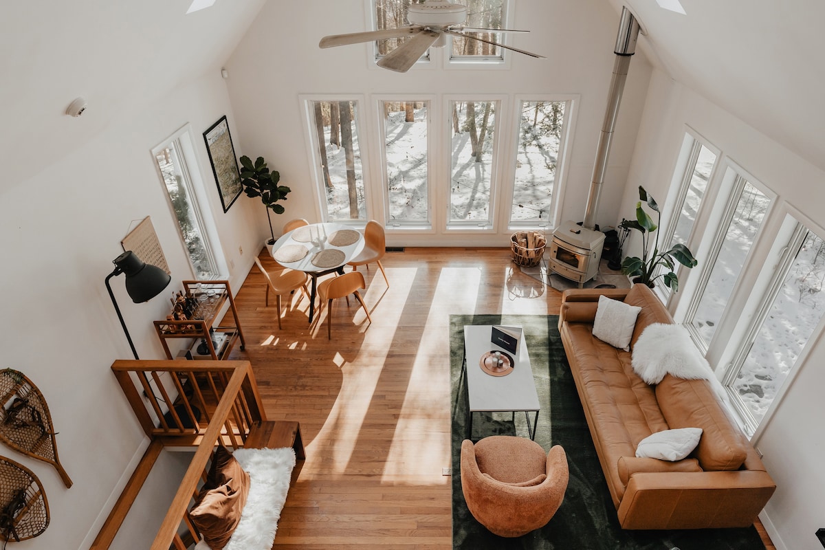 Overhead view of a sunlit, open-concept living space with vaulted ceilings, a ceiling fan, large windows overlooking a snowy forest, and a cozy seating area with a leather sofa.