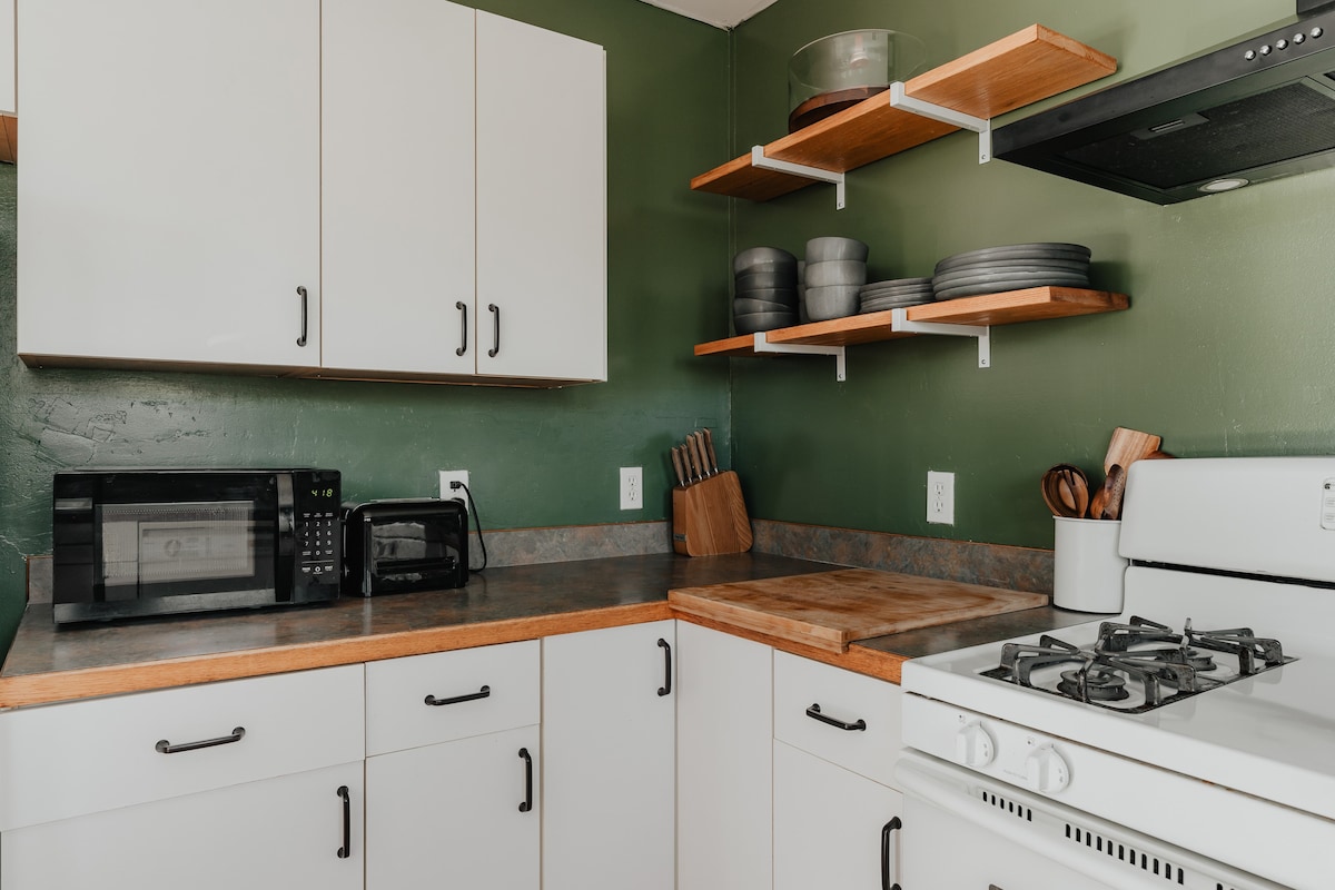 Another view of the kitchen showing white upper and lower cabinets, a white stove, and a microwave and toaster on the counter. Open wooden shelves display dishes, and a wooden cutting board is on the counter.