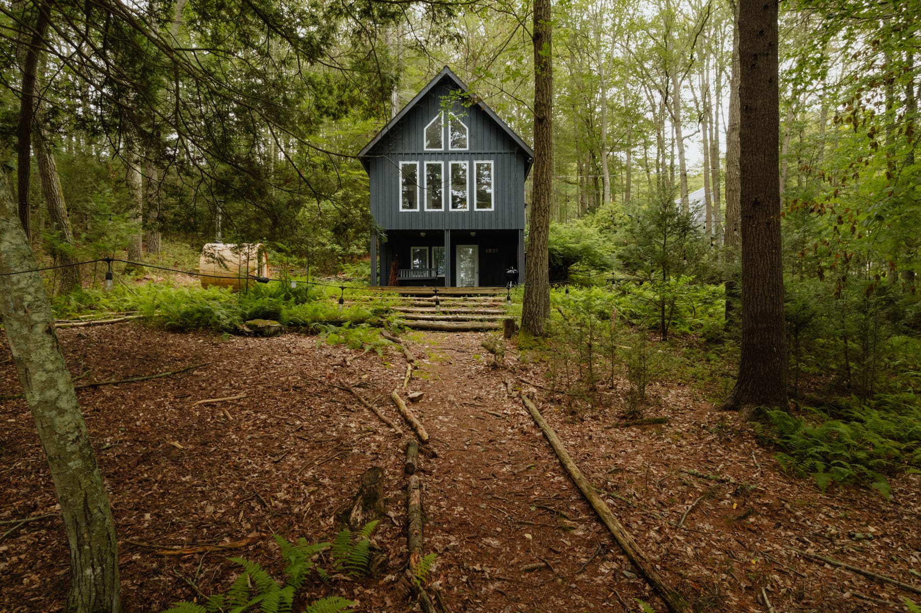 The front exterior of a dark gray cabin with white-framed windows, featuring a prominent A-frame style upper section, nestled in a dense forest with a natural path of wood steps leading to the entrance.
