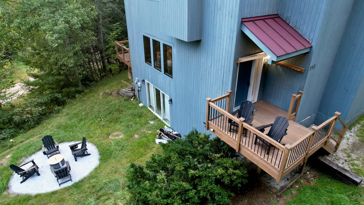 Ground-level view of the blue house's side, featuring a wooden deck with black Adirondack chairs, a sliding glass door, and a circular fire pit area on a white gravel patch in the yard.