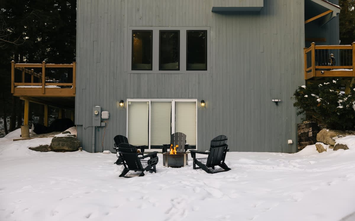Back exterior of the gray-blue cabin, featuring a patio area with four black Adirondack chairs arranged around a lit fire pit, all surrounded by snow. Large sliding glass doors lead into the house.