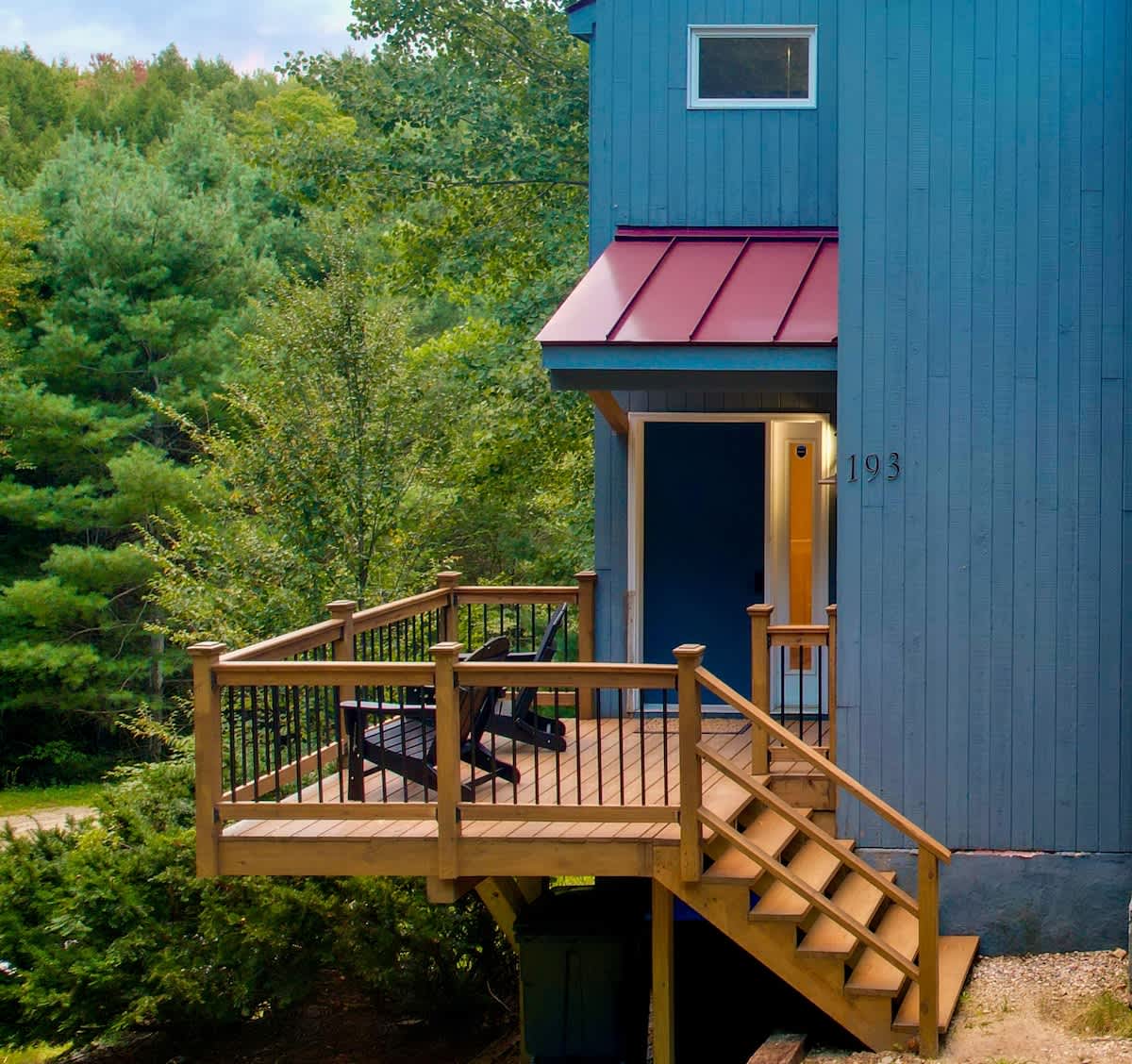 Detailed view of the blue house's front entrance, showcasing a raised wooden deck with black Adirondack chairs and sturdy stairs, leading to a door marked with the number '193'.