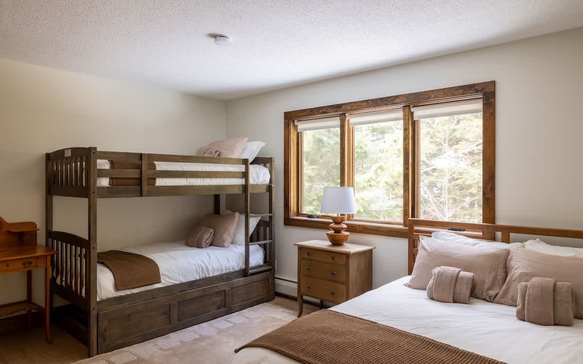 Cozy bedroom featuring a dark wood bunk bed with a trundle, alongside a queen-sized bed with light bedding. A wooden nightstand with a lamp sits between the beds, and large windows overlook a wooded area.